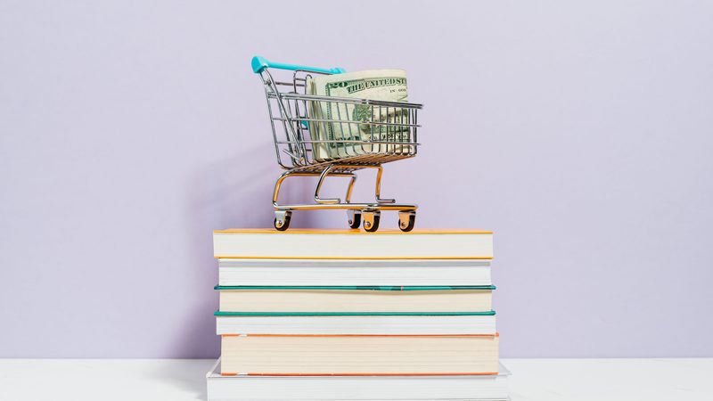 A miniature shopping cart with cash on a stack of books, representing selling books direct