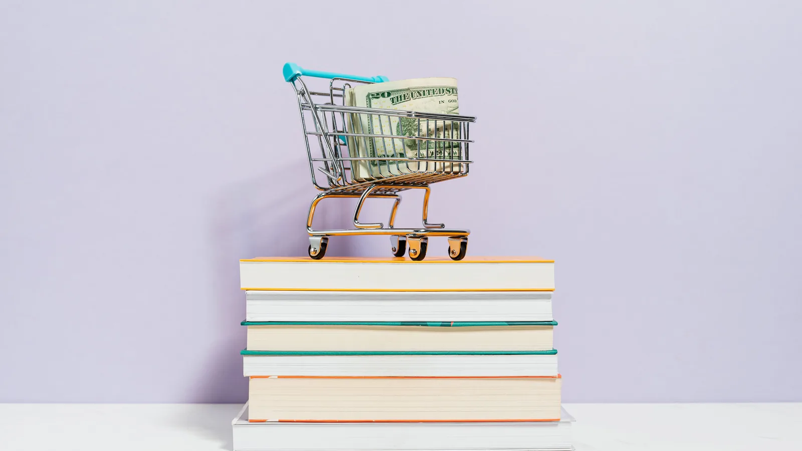 A miniature shopping cart with cash on a stack of books, representing selling books direct