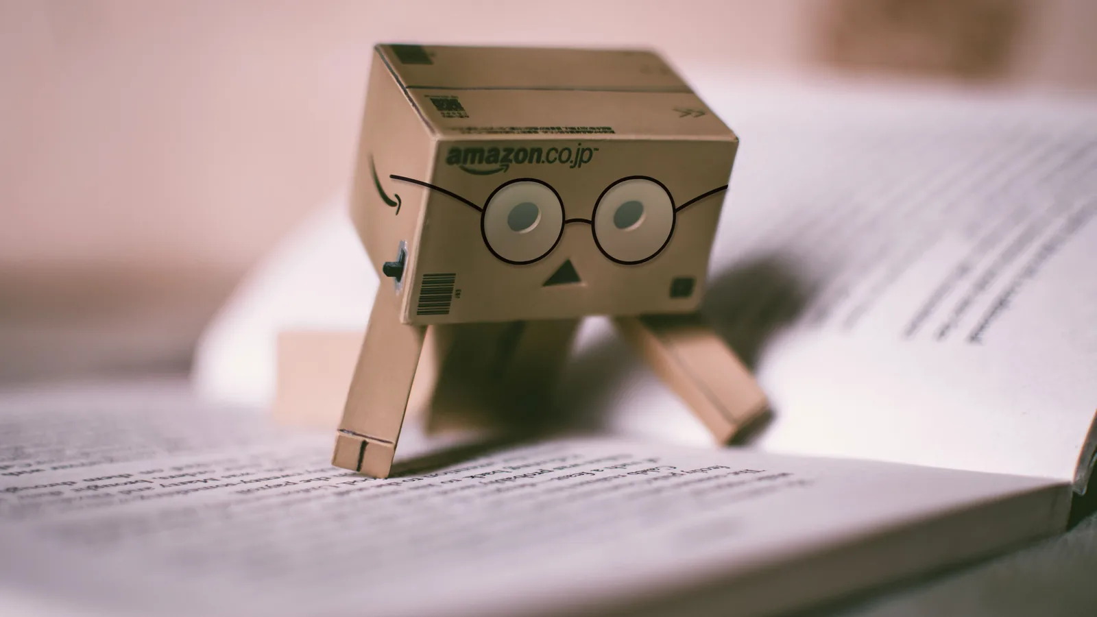 A small cardboard robot made from an Amazon box wearing glasses standing on an open book, representing AI and Amazon KDP